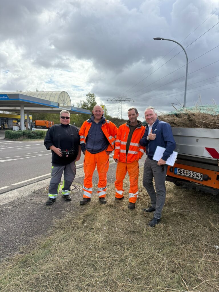 Heike von Komplex, zwei Mitarbeiter des Bauhofs Schönebeck und Thomas Kohl stehen am Ortseingang vor einem Anhänger mit Grünschnitt